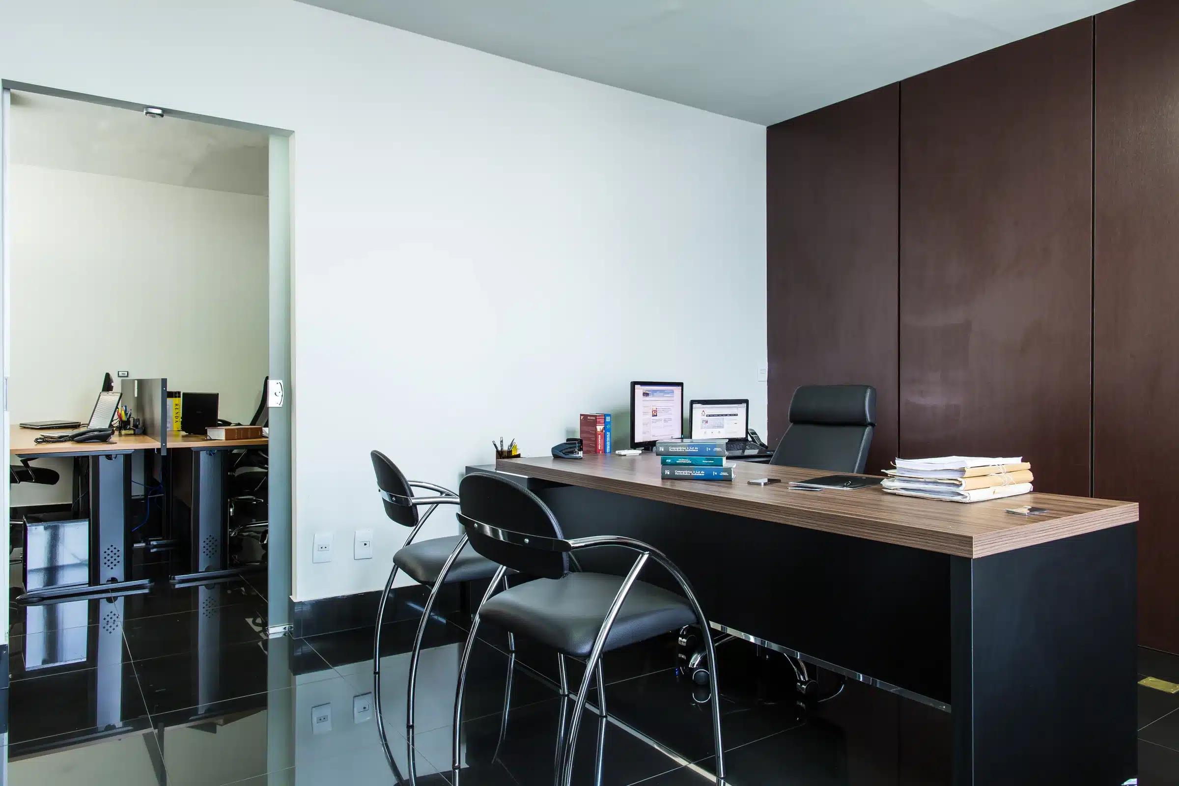 Sala de trabalho do escritório, com mesa executiva, parede de madeira escura e porta de vidro.